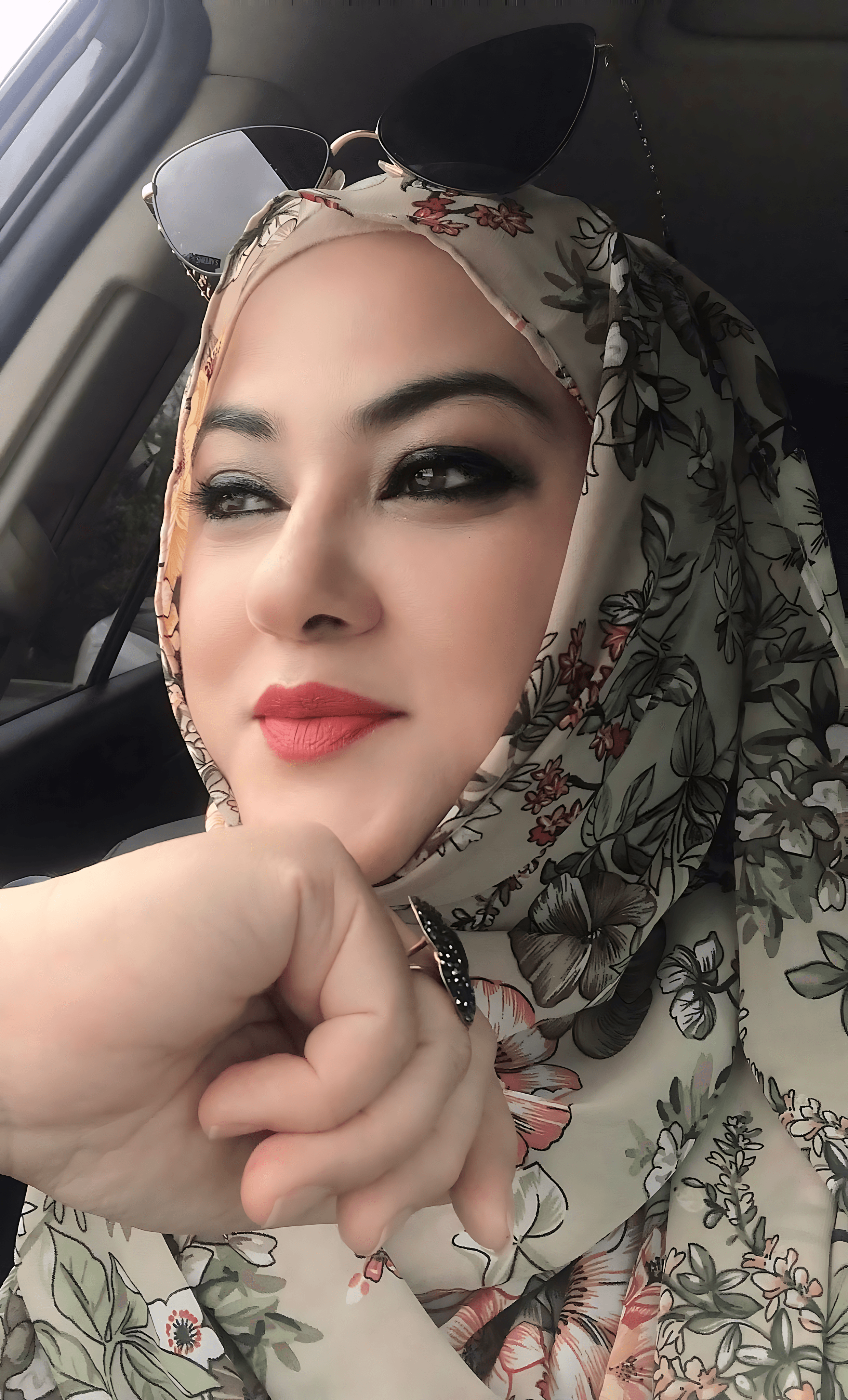 Woman wearing a floral hijab and sunglasses rests her chin on her hand inside a car.