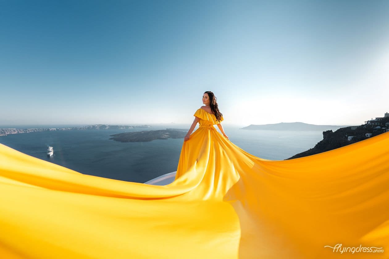 Woman in a vibrant yellow dress with a long, billowing train overlooking the Santorini coast.